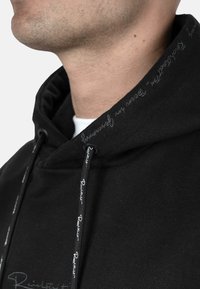 Felpa nera con dettagli a coulisse; presenta accenti di cucitura discreti e un tessuto morbido e texturizzato. L'apertura del collo è rotonda.