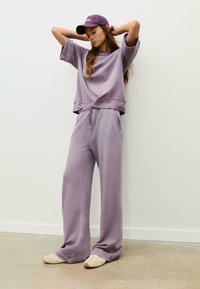 Fialové oversized mikina s krátkymi rukávmi a zodpovedajúce tepláky, obe vyrobené z mäkkej zmesovej bavlny. Celkový vzhľad dopĺňa fialová čiapka.