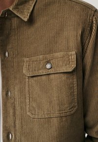 Brown corduroy shirt with a vertical ribbed texture, featuring a button-down collar, a single chest pocket, and a metal snap button detail.