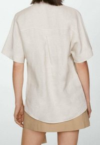 Chemise à manches courtes en lin beige avec un ourlet arrondi et un pli dans le dos. Texture lisse, design simple et coupe décontractée.