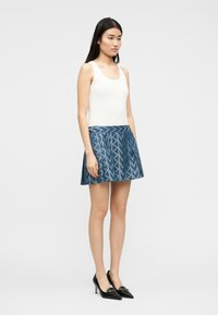 Γυναίκα που φοράει λευκό αμάνικο τοπ, μπλε φούστα με πτυχώσεις και μοτίβο, και μαύρες μυτερές γόβες, στέκεται μπροστά σε απλό φόντο.