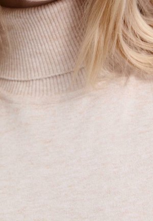 Pull col roulé beige côtelé porté par une personne aux cheveux blonds clairs visibles sur le côté droit.