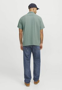 Krátký rukáv zelená košile se strukturovaným vzorem, spárovaná s modrými džíny a hnědou obuví. Námořnická čepice dokresluje celý outfit.