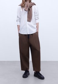 Camicia bianca a righe con le maniche arrotolate, pantaloni larghi marroni e sneakers nere. Un maglione di lana marrone poggiato attorno al collo.