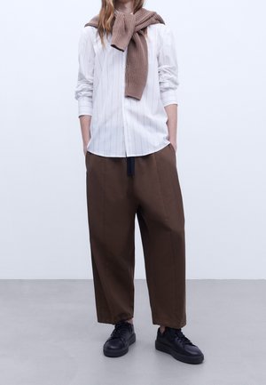 Chemise blanche à rayures avec des manches retroussées, pantalon large marron et baskets noires. Un pull en tricot marron drapé autour du cou.