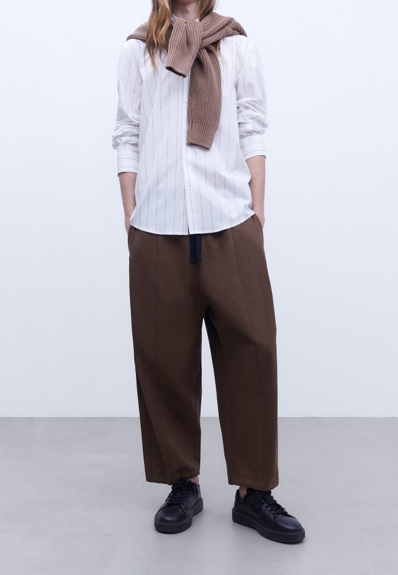 Camicia bianca a righe con le maniche arrotolate, pantaloni larghi marroni e sneakers nere. Un maglione di lana marrone poggiato attorno al collo.