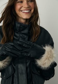 Sorte læderhandsker med en glat finish, prydet med beige kunstpelsdetaljer ved cuffen, parret med en sort læderjakke.