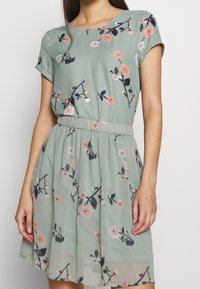 Blommig klänning i ljusgrön tyg med korta ärmar, samlad midja och blommönster i rosa och marinblått. Lätt och knälång design.