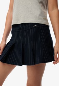 Gonna plissettata blu navy con strisce verticali bianche, caratterizzata da una vita piatta e un piccolo dettaglio metallico sul davanti.