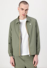 Jachetă ușoară de culoare verde măsline, cu fermoar frontal, manșete elastice și un accent în dungi multicolore de-a lungul fermoarului. Prezintă un logo pe piept.