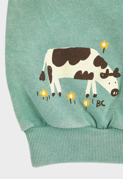 Tissu vert avec une illustration de vache marron et blanche broutant parmi de petites fleurs jaunes, avec les initiales "BC" près de la vache.
