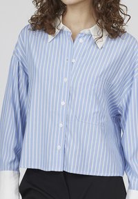 Lysblå skjorte med hvide lodrette striber, blødt stof, klassisk krave og en enkelt brystlomme. Lange ærmer med hvide manchetter.