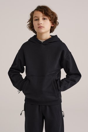 Lapsi, jolla on lyhyet kiharat hiukset, pukeutuneena mustaan huppariin ja mustiin housuihin, kädet hupun taskuissa, neutraali tausta.