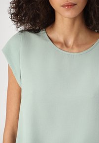 Blouse vert clair avec un col rond, des manches courtes et un tissu à texture fine, offrant une finition lisse et un design simple.