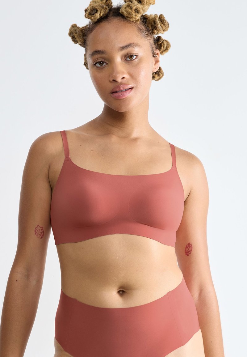 Femme aux cheveux texturés portant un soutien-gorge sans coutures et une culotte taille haute assortis de couleur rouille, debout devant un fond clair uni.