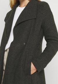 Cappotto in misto lana verde scuro con un ampio colletto, chiusura doppiopetto, tasche laterali e tessuto con texture.