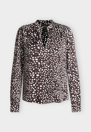 Mujer con un conjunto negro suelto con puntos blancos irregulares, sosteniendo un bolso blanco en la mano izquierda, de pie frente a un fondo claro liso.