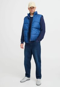 Zila pūku veste ar augstu apkakli, tumši zila garā piedurkne krekls zem tās, tumši džinsi un baltas sporta apavi; bēšs cepure ar logotipu.