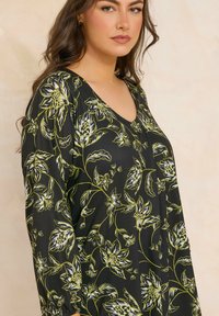 Blusa nera con motivo floreale verde, scollatura a V, maniche lunghe e polsini elasticizzati. Tessuto morbido e liscio con dettagli floreali intricati.