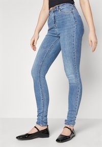 Jeans de denim azul ajustados con cintura alta, que presentan un diseño de cinco bolsillos y un ligero desgaste. Combinados con zapatos planos negros.