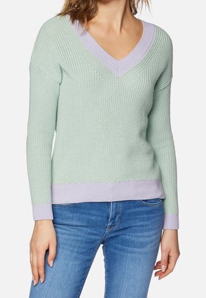Femme portant un pull en maille vert clair à col en V avec une bordure lavande, associé à un jean bleu, debout devant un fond uni.