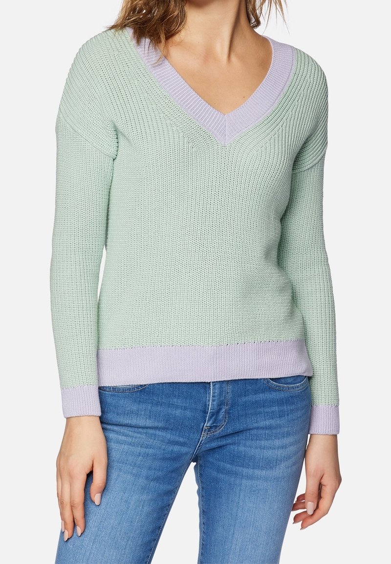 Femme portant un pull en maille vert clair à col en V avec une bordure lavande, associé à un jean bleu, debout devant un fond uni.