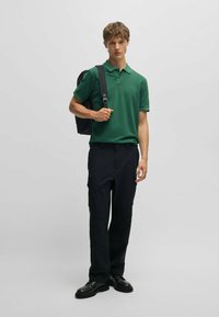 Grön polo skjorta i kombination med svarta cargo-byxor. Modellen bär en svart ryggsäck och har på sig svarta skor. Enkel bakgrund.