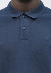 Gros plan sur une personne portant un polo bleu foncé avec deux boutons fermés et un petit logo brodé sur la poitrine.