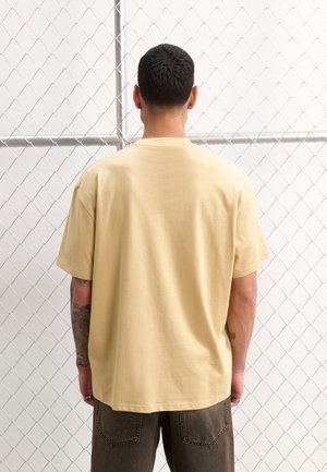 Homme aux cheveux courts et foncés, portant un t-shirt beige uni et un pantalon foncé, debout face à une clôture en grillage.