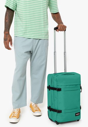 Green fabric suitcase with black straps, two front pockets, retractable handle, and wheels, positioned next to a person in light clothing.