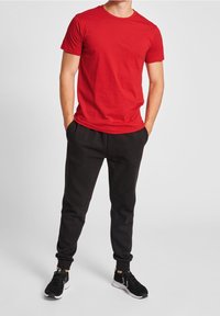 Rotes Baumwoll-T-Shirt mit kurzen Ärmeln, kombiniert mit schwarzen Sweatpants mit elastischen Bündchen. Schwarze Sportschuhe runden das Outfit ab.