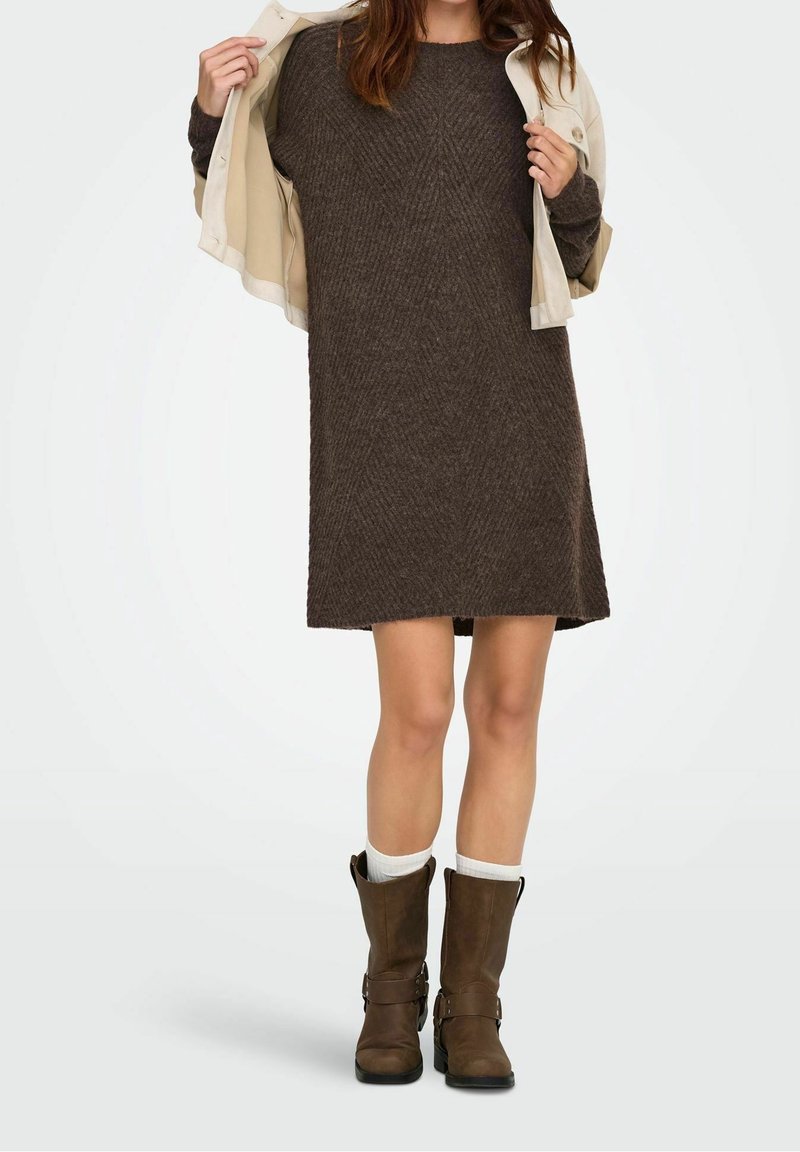 Femme portant une robe en maille texturée marron, des bottes marron mi-mollet, des chaussettes blanches, et tenant une veste beige ouverte sur les épaules.