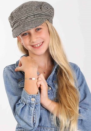Casquette en pied-de-poule noir et blanc, veste en denim, bracelet argenté avec un détail rouge, cheveux blonds longs, visage souriant, mains reposant sur le poignet.