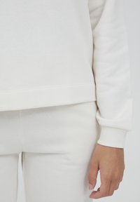 Felpa bianca con polsini a coste e orlo dritto, abbinata a pantaloni bianchi coordinati. Tessuto in cotone morbido con texture liscia.