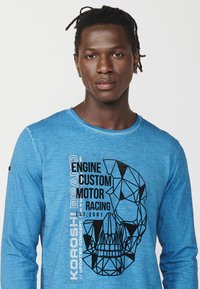 Blue long-sleeve shirt made of soft fabric featuring a black geometric skull graphic with text: "ENGINE CUSTOM MOTOR RACING EST. 2001."