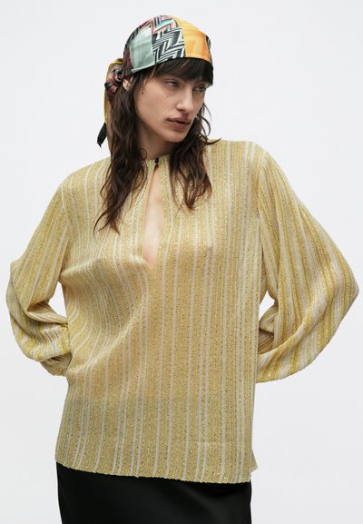 Gul- og guldbenet bluse med dyb V-hals og lange ærmer, med en diskret glans og struktureret stof. Rundet kantdetalje.