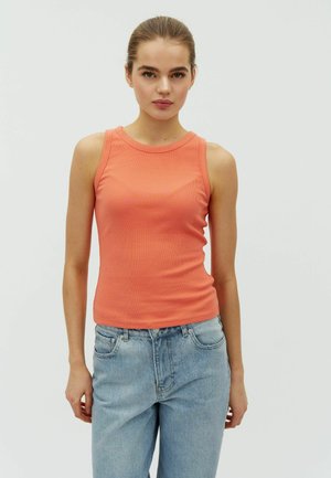 Mladá žena v koralovom tielku bez rukávov a svetlo modrých džínsoch, stojaca na jednoduchom svetlom pozadí.