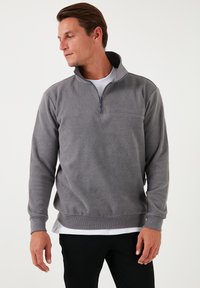 Pullover grigio in pile con colletto alto, chiusura con zip a metà, polsini a coste e vita aderente, indossato sopra uno strato bianco.