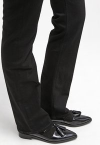 Pantalon noir droit porté avec des mocassins en cuir noir brillant ornés de détails à pompons sur fond blanc uni.