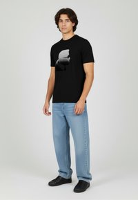 Czarny T-shirt z graficznym profilem w odcieniach szarości, zestawiony z jasnoniebieskimi dżinsami i czarnymi trampkami. Casualowy krój i styl.