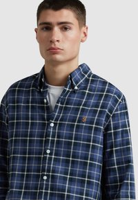 Camicia a quadri blu e bianca con colletto button-down, caratterizzata da un sottile logo arancione. Realizzata in un morbido tessuto di cotone con una texture liscia.