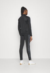 Čierny teplákový komplet so zipsom a slim fit nohavicami. Bunda má golier, nohavice majú bočné pruhy z reflexného materiálu. Biele tenisky.
