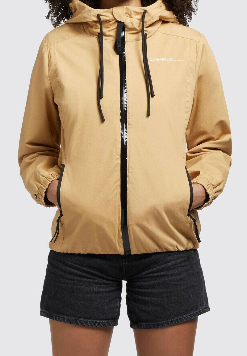 Persona con chaqueta con capucha color beige con cremallera negra y cordones, manos en los bolsillos, combinada con pantalones cortos de mezclilla negros.