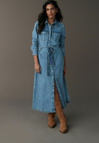 Robe chemise en denim à manches longues, avec un devant à boutons, une taille ceinturée et une fente sur le côté, en délavé bleu clair, comportant deux poches poitrine.