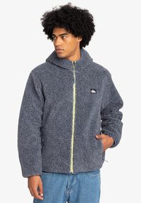 Veste en polaire grise avec un col montant, dotée d'une fermeture éclair jaune et d'un logo brodé sur la poitrine gauche ; elle a deux poches latérales.