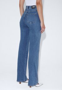 Jeans in denim blu a vita alta con taglio dritto, dotati di una tasca posteriore e di un'etichetta con il marchio sulla cintura. Texture liscia e cuciture minime.