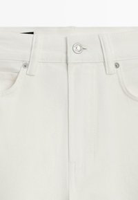 Détail avant d'un jean en denim blanc montrant le bouton, la fermeture éclair, les passants de ceinture et les poches partielles.