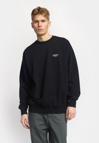 Sudadera negra de algodón con cuello redondo. Presenta un pequeño logotipo blanco en el pecho. Mangas largas con puños y bajo acanalados.