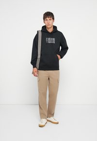 Μαύρη κουκούλα με γραφικά "VISION", ζεστό βαμβακερό υλικό, τσέπη καγκουρό. Μπεζ cargo παντελόνι με πλαϊνές τσέπες. Λευκά αθλητικά παπούτσια ολοκληρώνουν την εμφάνιση.