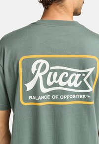 Vihreä puuvillapaita, jossa on iso valkoinen ja keltainen logo selässä, jossa lukee "Ruca" ja "BALANCE OF OPPOSITES™."